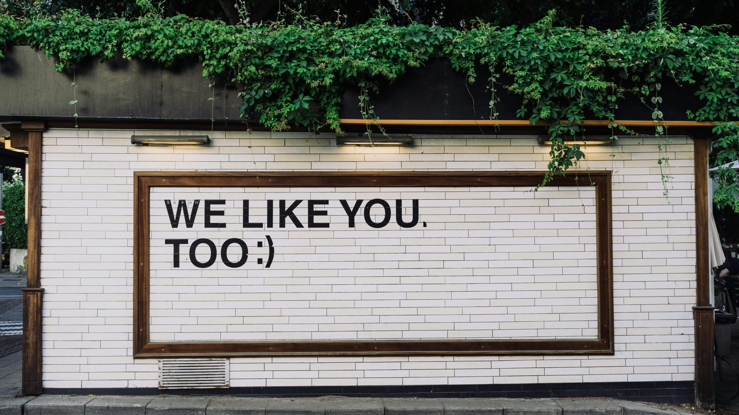 unreasonable hospitality- Blog image for Anton Tonev's (Rexera Co-founder) LinkedIn Newsletter- while wall with greenary, sign saying we like you too